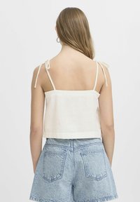 Top beige recortado con tirantes finos, que presenta un escote cuadrado y una textura suave, combinado con pantalones cortos de mezclilla azules.
