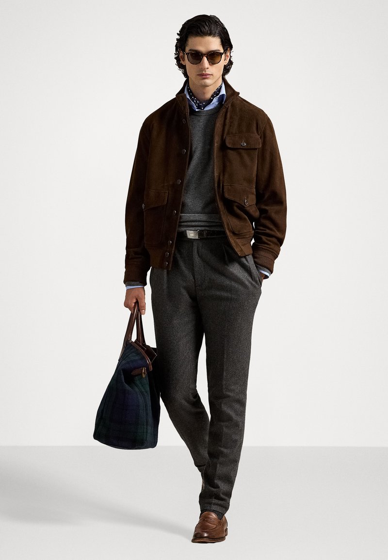 Bruine suède jas, grijze trui, gestreept blauw shirt, grijze broek, bruine riem, bruine schoenen, houdt een geruite tas vast. Donkere zonnebril.