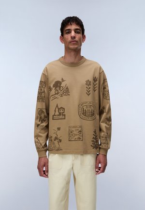Mand står mod en ensfarvet baggrund iført en lysebrun sweatshirt med naturinspirerede motiver og offwhite bukser, vendt fremad.