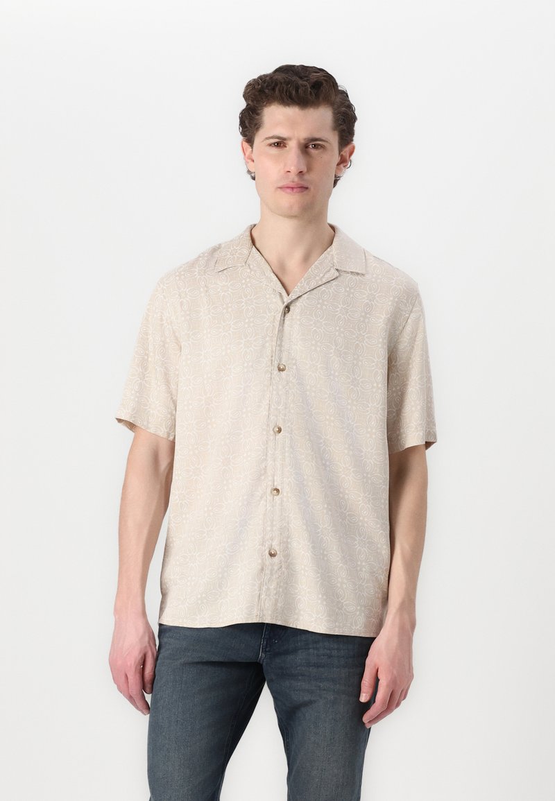 Camisa de manga corta beige con un sutil patrón circular, con botones en el frente y un cuello relajado, combinada con jeans oscuros.