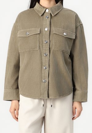 Femme portant une chemise boutonnée en velours côtelé marron clair avec deux poches poitrine et un pantalon de couleur crème.