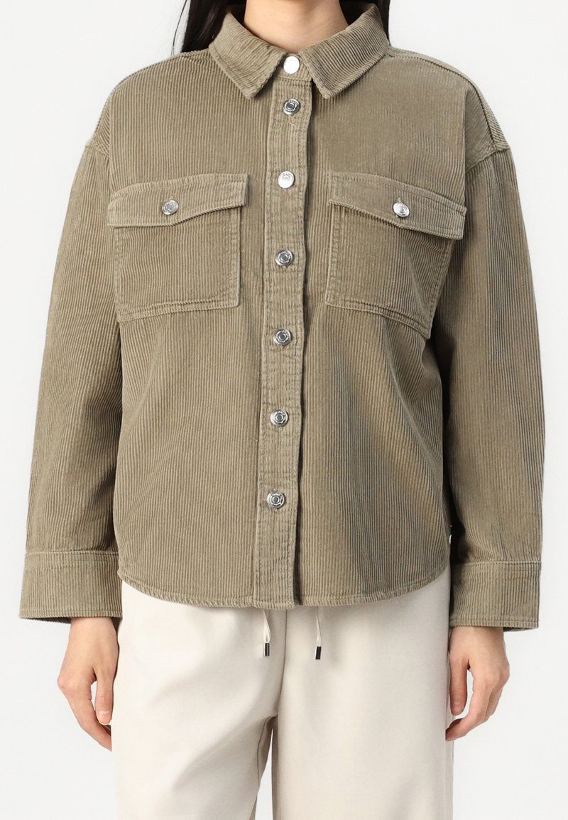 Femme portant une chemise boutonnée en velours côtelé marron clair avec deux poches poitrine et un pantalon de couleur crème.