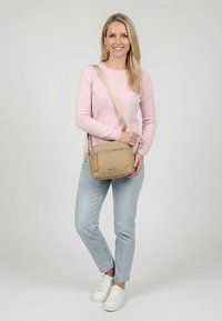 Frau steht lächelnd, trägt einen rosa Pullover, hellblaue Jeans, weiße Sneakers und hält eine hellbraune Umhängetasche mit Reißverschlusstasche vorne.