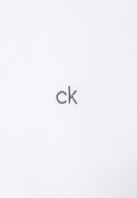 Bílý polo tričko s texturovanou tkaninou a vyšitým šedým logem "ck" na levé straně hrudi.