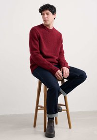 Punainen tekstuurilla kudottu villapaita, jossa on pyöreä kaula-aukko, yhdistettynä tummiin farkkuihin ja ruskeisiin nilkkasaappaisiin. Malli istuu puuistuimella.