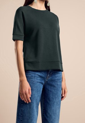 Frau trägt ein dunkelgrünes kurzärmeliges Oberteil und blaue Denim-Jeans, mit langen Haaren und großen durchsichtigen Ohrringen, steht vor einem einfarbigen Hintergrund.