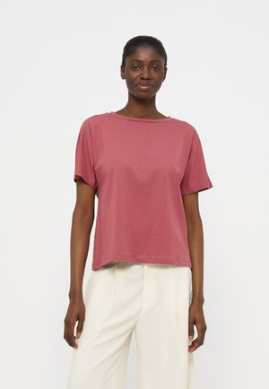 Γυναίκα με σκούρο δέρμα και μαλλιά δεμένα πίσω, που φοράει μια κοντομάνικη μπλούζα σε ξεθωριασμένο ρόζ και άνετο λευκό παντελόνι, στέκεται μπροστά σε λευκό φόντο.