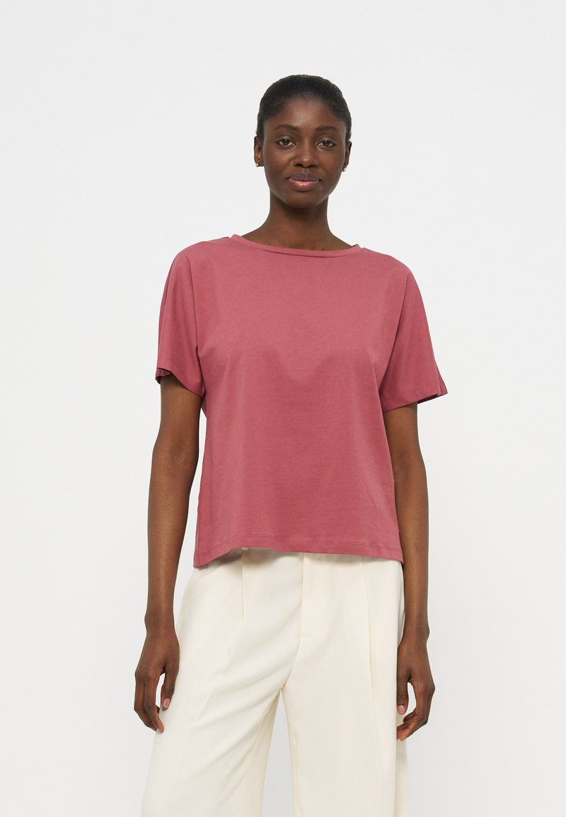 Γυναίκα με σκούρο δέρμα και μαλλιά δεμένα πίσω, που φοράει μια κοντομάνικη μπλούζα σε ξεθωριασμένο ρόζ και άνετο λευκό παντελόνι, στέκεται μπροστά σε λευκό φόντο.