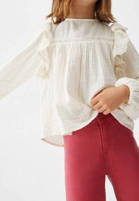 Vit bomullskjorta med volangärmar och spetsdetaljer, kombinerad med figurnära röda byxor. Texturerat tyg med subtila detaljer och en något lös passform.