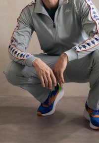 Homme accroupi portant un survêtement gris clair avec des bandes de logo bleues et blanches sur les manches et des baskets bleues, orange et vertes.