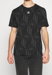 Man som bär en svart Adidas T-shirt med mörkt geometriskt mönster och vitt Adidas-logotyp på bröstet, kombinerat med mörka jeans.