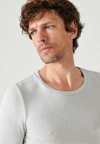 Maglietta grigia chiara a maniche corte con scollatura rotonda, texture liscia e design aderente, presentata su uno sfondo neutro.