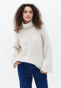 Cremfarbener Rollkragenpullover aus weichem Stoff, mit weitem Ärmel und geripptem Saum. Getragen mit blauen Cordhosen.