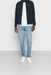 Námořnická lehká zipová bunda přes bílý tričko, spárovaná se světle modrými džíny a bílými teniskami. Jednoduchý design s žebrovanými manžetami.