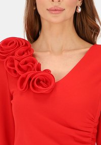 Czerwona sukienka z dekoltem w serek, miękkim materiałem oraz trójwymiarowymi różami z tkaniny na lewym ramieniu. Teksturowane detale podkreślają wizualne zainteresowanie.