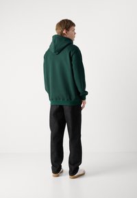 Sudadera verde oscura con un corte relajado y capucha grande, combinada con unos vaqueros negros holgados y zapatillas blancas, vista desde la espalda.