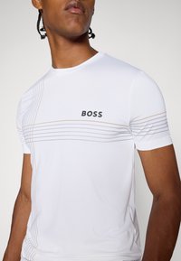 T-shirt sportivo bianco realizzato in tessuto morbido, con accentuazioni lineari grigie e dorate, e logo "BOSS" sulla parte frontale in caratteri neri in grassetto.