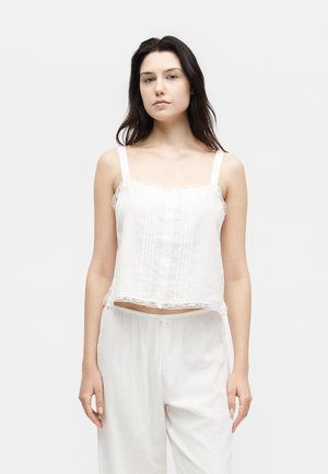 Jonge vrouw met donker haar, gekleed in een witte mouwloze top met kanten rand en losse witte broek, staande tegen een effen achtergrond.