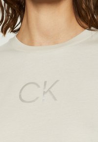 T-shirt beige chiaro realizzato in tessuto morbido, con stampa del logo metallico "CK" sul davanti e un design classico a girocollo.
