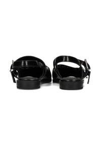 Sandali bassi in vernice nera con cinturini alla caviglia e fibbie argento, visti da dietro su sfondo bianco.
