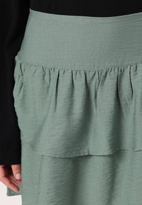 Grønn lagdelt skjørt laget av lett stoff, med en volangdesign. Livet er glatt, noe som står i kontrast til den teksturerte kroppen.