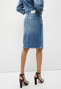 Falda lápiz de mezclilla en azul claro. Presenta bolsillos traseros y costuras visibles. Se lleva con sandalias de tacón negro con detalles decorativos.