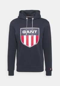 Sudadera con capucha azul marino con un gran logo en forma de escudo que incluye el texto "GANT" y detalles a rayas rojas y blancas. Cuenta con cordones ajustables y mangas con puños.