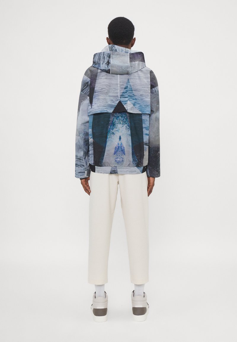 Kapuzenshirt mit einem abstrakten Blau- und Graumuster. Hergestellt aus leichtem Material mit lockerer Passform und elastischen Bündchen, kombiniert mit cremefarbenen Hosen.