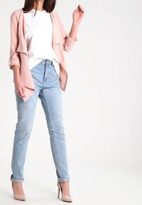 Woman wearing light pink blazer, white top, light blue jeans, and beige high heels standing against white background.