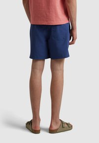 Pantaloni da bagno blu con un design semplice, tasca laterale e tessuto leggero, abbinati a sandali marroni con dettaglio a fiocco.