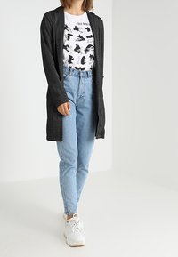 Long gilet noir porté sur un t-shirt blanc à motif avec des silhouettes d'animaux noires, associé à un jean bleu clair et des baskets blanches.