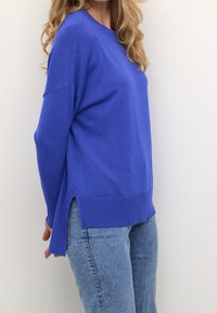 Blauer gestrickter Pullover mit lockerem Schnitt, geripptem Halsauschnitt und seitlichen Schlitzen. Kombiniert mit hellblauen, eng anliegenden Jeans.