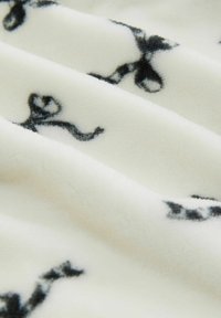 Fleece-tyg med en krämfärgad bakgrund och svarta snöringsmönster, mjuk textur och subtil lyster, som visar mjuka veck och lager.