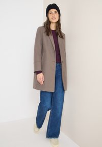 Vaaleanruskea villatakki, jossa on muotoiltu malli, kerrostettuna purppuranvärisen neuleen ja leveälahkeisten sinisten farkkujen kanssa, yhdistettynä kermaisiin lenkkitossuihin.