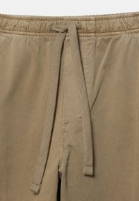 Bukser med elastisk talje i beige stof med matchende snor bundet i en sløjfe foran i midten.