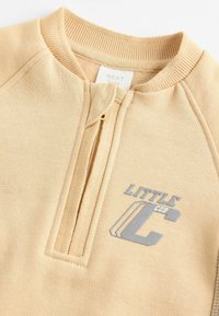 Helles beiges Sweatshirt mit einem Reißverschlusskragen, das eine weiche Textur aufweist. Graues "LITTLE CUB"-Logo und geometrische Akzente sind vorhanden.