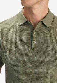 Olivinvihreä teksturoitu poolopaita, jossa on kolmen napin suljinta ja lyhyet hihat. Paidassa on hienovarainen geometrinen kuvio ja pehmeä kangas.