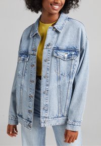 Ljusblå denimjacka med två bröstfickor, silverknappar och en avslappnad passform. Tyget har en lätt blekning och synliga sömmar.