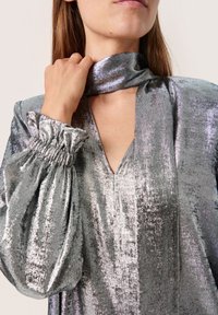 Blusa metálica plateada con escote en V, mangas abullonadas y puños recogidos. Textura suave con un acabado brillante, creando una apariencia reflectante.