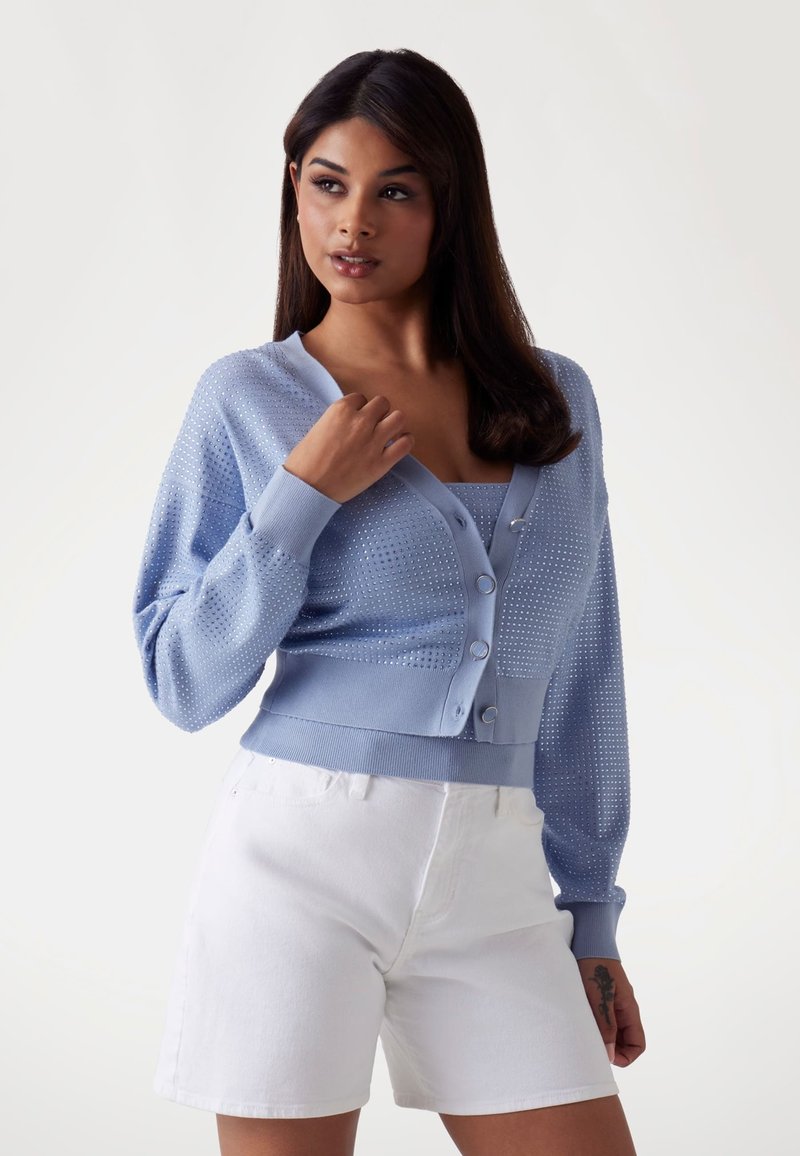 Cardigan azzurro chiaro con texture a pois, scollo rotondo e chiusura con bottoni. Indossato sopra un top aderente abbinato e abbinato a pantaloni corti bianchi.
