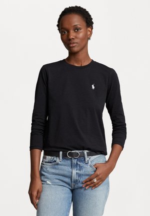 Svart langermet t-skjorte laget av bomull, med en liten hvit logo på brystet. Kombinert med slitte lyse blå denimjeans.