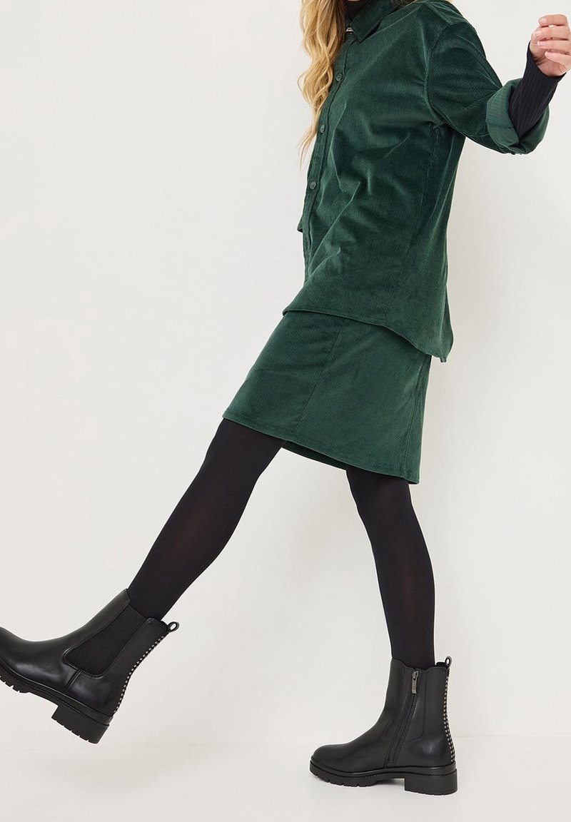 Femme portant une jupe en velours côtelé vert foncé et une chemise, des collants noirs et des bottines en cuir noir, levant la jambe gauche vers l'avant en marchant.