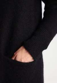 Černý, měkký pletený svetr s kapsou a ribováním na manžetách, detail na ruku odpočívající v kapse s chlupatou texturou.