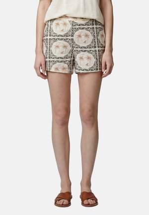 Frau trägt beige Shorts mit Palmen- und Segelboot-Motiv, weißes Top und braune Sandalen, steht vor weißem Hintergrund.