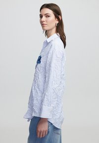 Camicia a righe blu e bianche con bottoni, caratterizzata da una texture stropicciata e un dettaglio floreale in tessuto blu sul petto. Maniche lunghe con polsini.