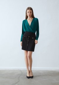 Türkise Wickelbluse mit langen Ärmeln, kombiniert mit einem schwarzen, engen Minirock mit goldenen Knöpfen und Taschen. Glatter Stoff mit einer strukturierten Form.