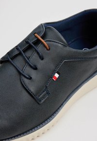 Zapato de piel marino con textura perforada, detalle de cordones marrones y suela blanca. Presenta un pequeño logo tricolor en el lateral.