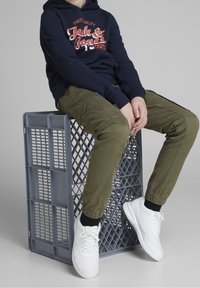 Granatowy hoodie z logo "Jack & Jones", oliwkowe spodnie cargo z elastycznymi mankietami oraz białe trampki na szarym plastikowym pudle.