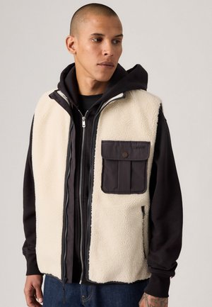 Gilet en polaire crème avec une poche en tissu foncé. Il dispose d'un zip à l'avant, d'une capuche noire en dessous et d'une coupe décontractée.