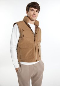 Braune Steppweste mit hohem Kragen, Frontreißverschluss und vier gro�ßen Taschen. Über einem weißen Langarmshirt getragen und mit hellgrauen Hosen kombiniert.